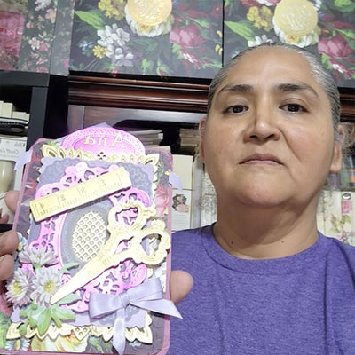 A person holding a decorative handmade card featuring scissors, a ruler, and floral designs. The person is wearing a purple shirt and standing in front of floral-patterned boxes.