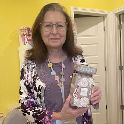 A woman with glasses, wearing a floral jacket and a colorful necklace, holds a decorated box in a room with a yellow wall and white doors.