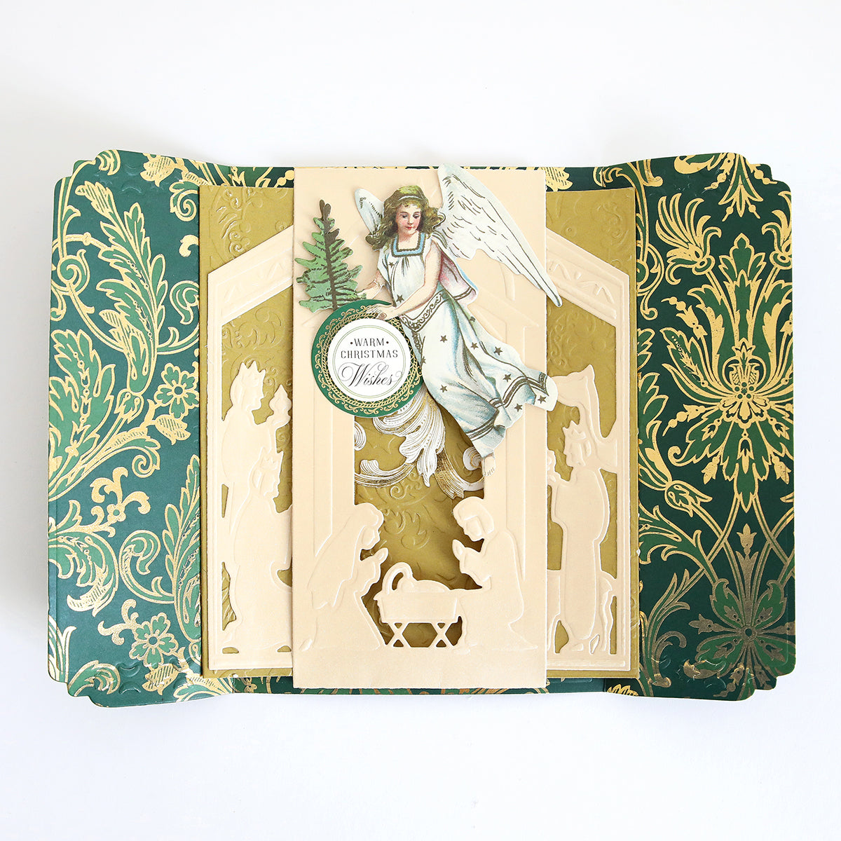 A decorative Christmas card created with the Nativity Scenes Die Set Collection, featuring a Nativity shadow box die-cut scene, a flying angel holding a tree, and the text “Warm Christmas Wishes.”