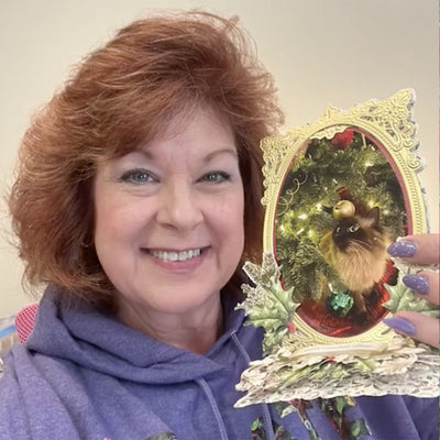 A woman with red hair holds a Christmas card featuring a cat in front of a Christmas tree. She is smiling and wearing a purple hoodie.