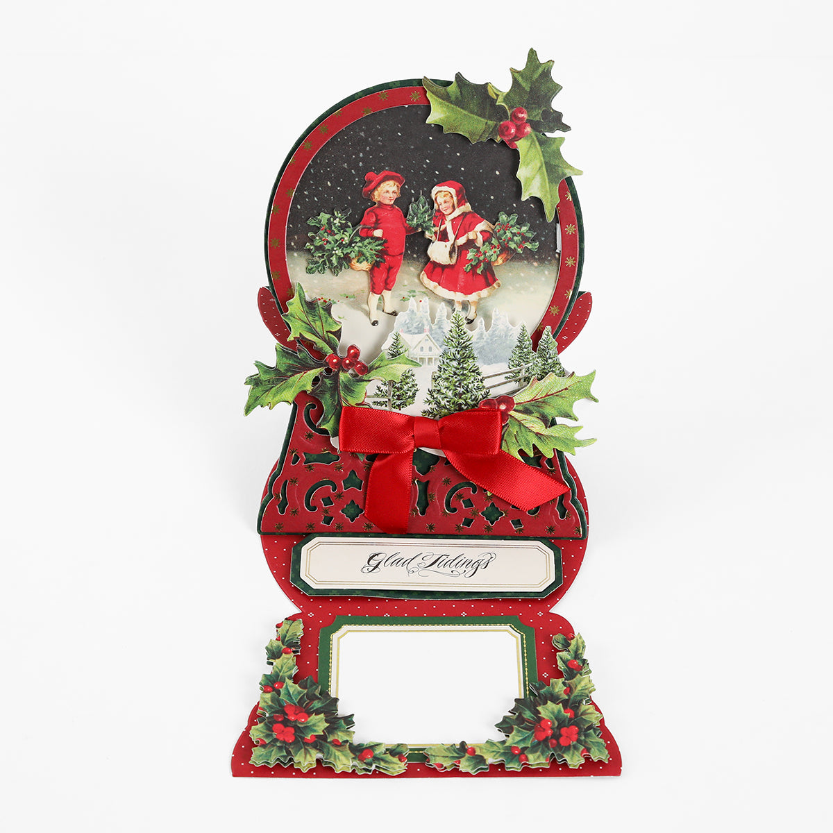 A festive holiday card adorned with children dressed in winter attire, holly leaves, a red bow, and the phrase “Glad Tidings” on a crisp white background—an ideal choice for creative projects or pairing with your Snowglobe Easel Card Dies.