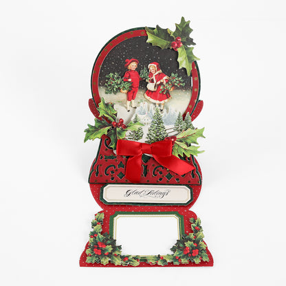 A festive holiday card adorned with children dressed in winter attire, holly leaves, a red bow, and the phrase “Glad Tidings” on a crisp white background—an ideal choice for creative projects or pairing with your Snowglobe Easel Card Dies.