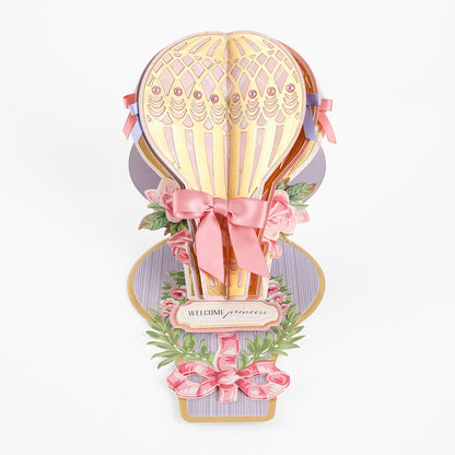 Create standout handmade cards with the Hot Air Balloon Easel Die—a 3D pop-up card featuring a gold and pink hot air balloon, beautifully accented with ribbons, flowers, and a “Welcome princess” sign on a decorative pastel background.