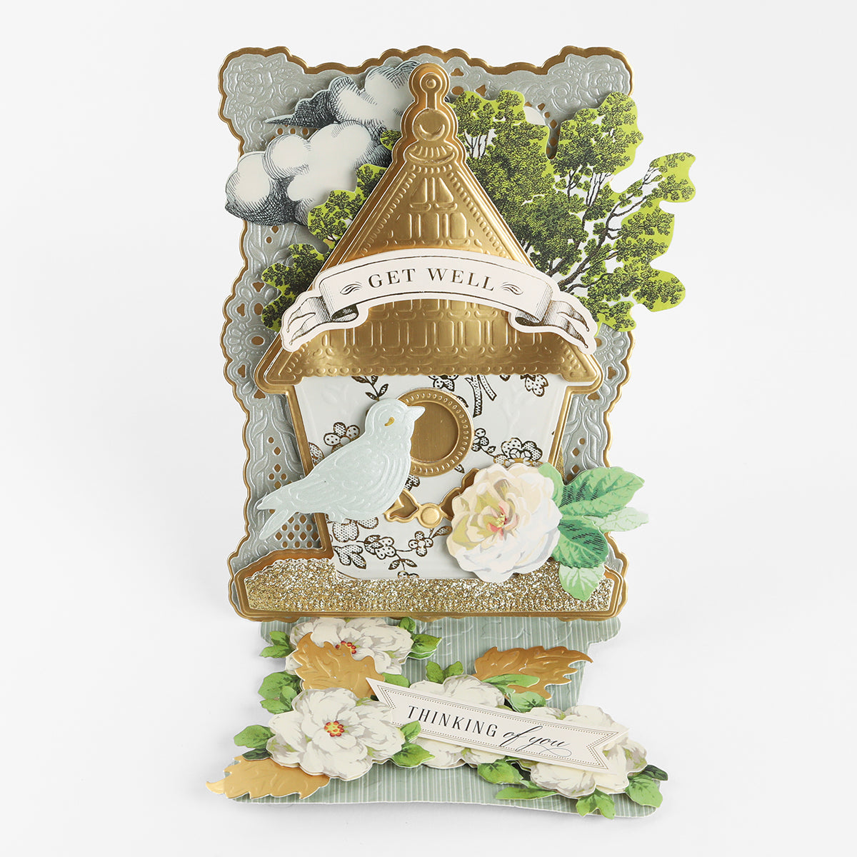 Send warm wishes with a handcrafted card made using the Birdhouse Easel Card Dies—featuring a sparkling gold birdhouse, an adorable blue bird, white and yellow blossoms, lush greenery, and cheerful “GET WELL” and “THINKING of you” banners. Perfect for thoughtful get-well messages or as a charming housewarming greeting.