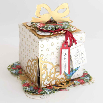 An elegant Explosion House Dies and Materials gift box features gold accents, floral patterns, a gold bow, die-cut embellishments, and tags that read “You make my heart smile” and “Your friend.”.