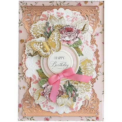 A detailed, floral-themed Wildflower Meadow Papercrafting Kit features intricate gold and pink designs with embellishments, a decorative butterfly, and a pink ribbon, with "Happy Birthday" inscribed at the center.