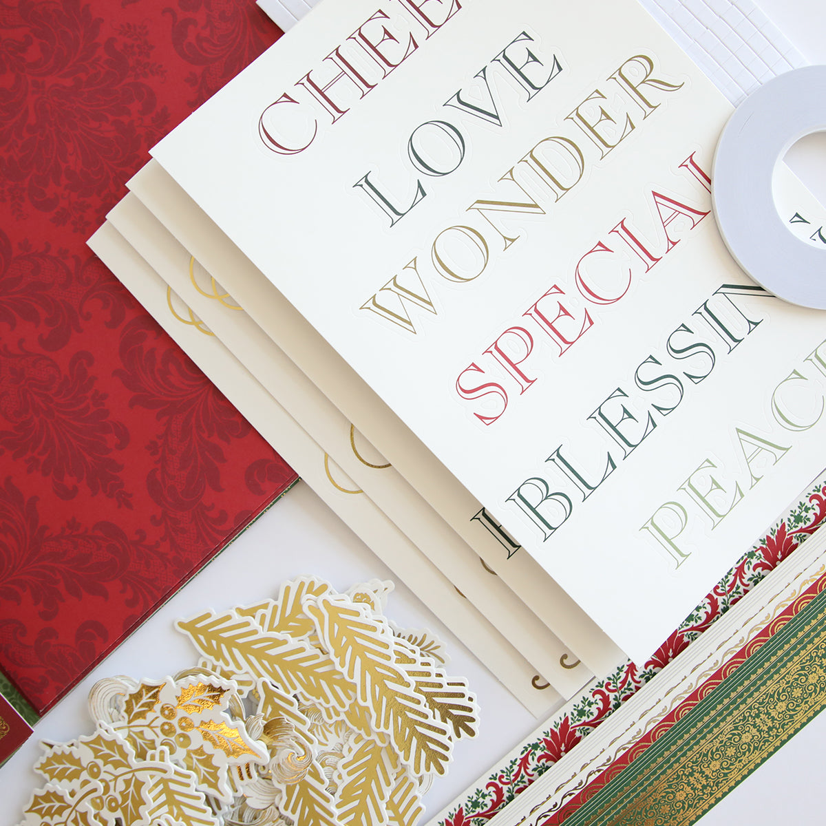 A set of Simply Noel Papers and Embellishments including double-sided cardstock with words like "LOVE" and "PEACE," decorative gold cutouts, a red and gold ribbon, and a white roll of tape.