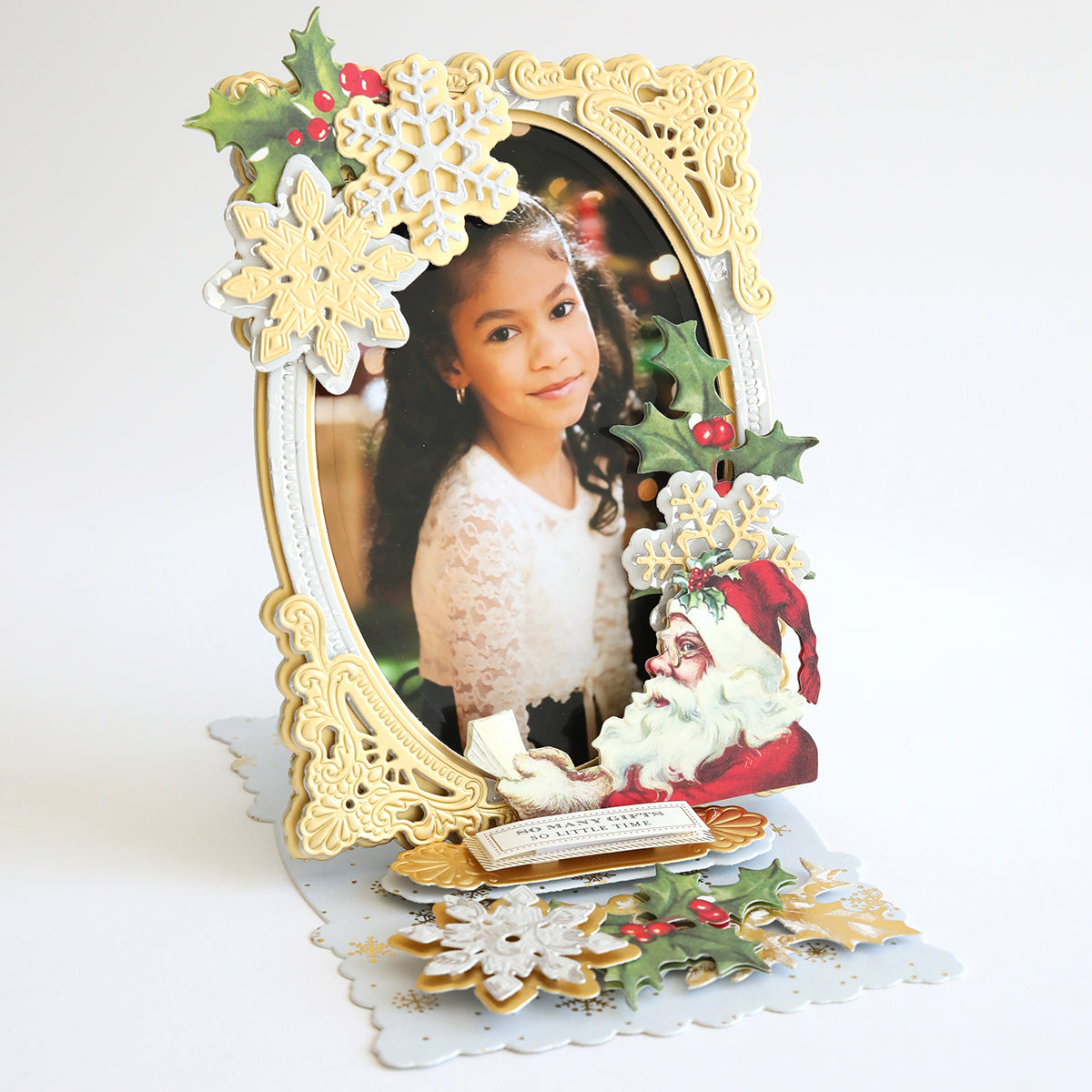 Decorative holiday-themed photo frame crafted from Christmas Double Sided 300gsm Cardstock, featuring an ornate gold oval border adorned with snowflakes, holly leaves, and a Santa Claus illustration at the bottom right corner. Photo of a young girl inside. AG Members exclusive design!