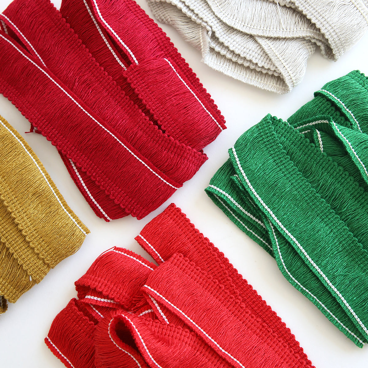 Various colored fabric trims, including red, green, gold, and cream—reminiscent of holiday-inspired colors—are neatly arranged on a white surface. Christmas Perfect Palette Fringe is prominently featured among them.