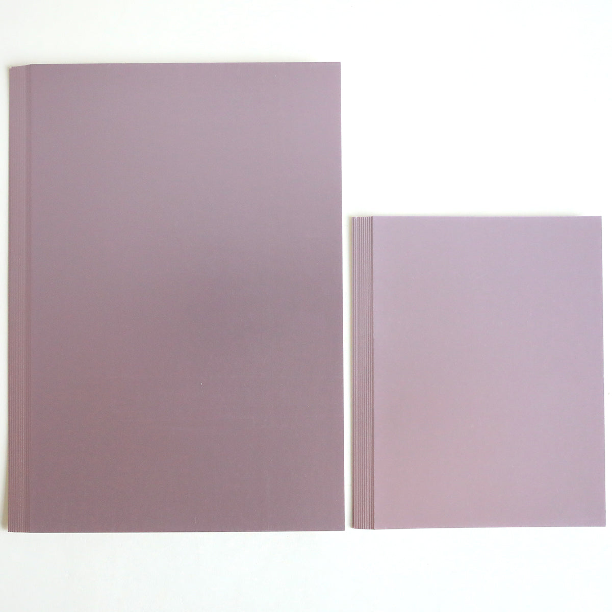 A set of three luxury matte foil cardstock notebooks—in lavender, blue, and green—rests on a white surface; one large notebook lies beside the neatly stacked trio.