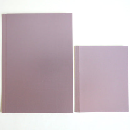 A set of three luxury matte foil cardstock notebooks—in lavender, blue, and green—rests on a white surface; one large notebook lies beside the neatly stacked trio.