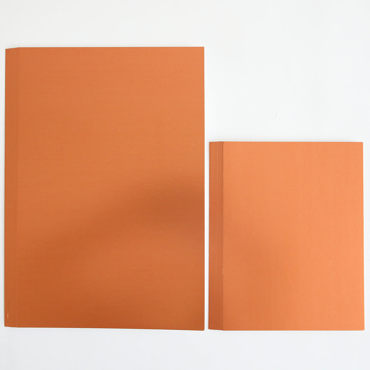 Two closed orange books are placed side by side on a white surface; the book on the left is larger than the one on the right, resembling stacked sheets from the Fall Matte Foil Cardstock Bundle—perfect for your next creative project.