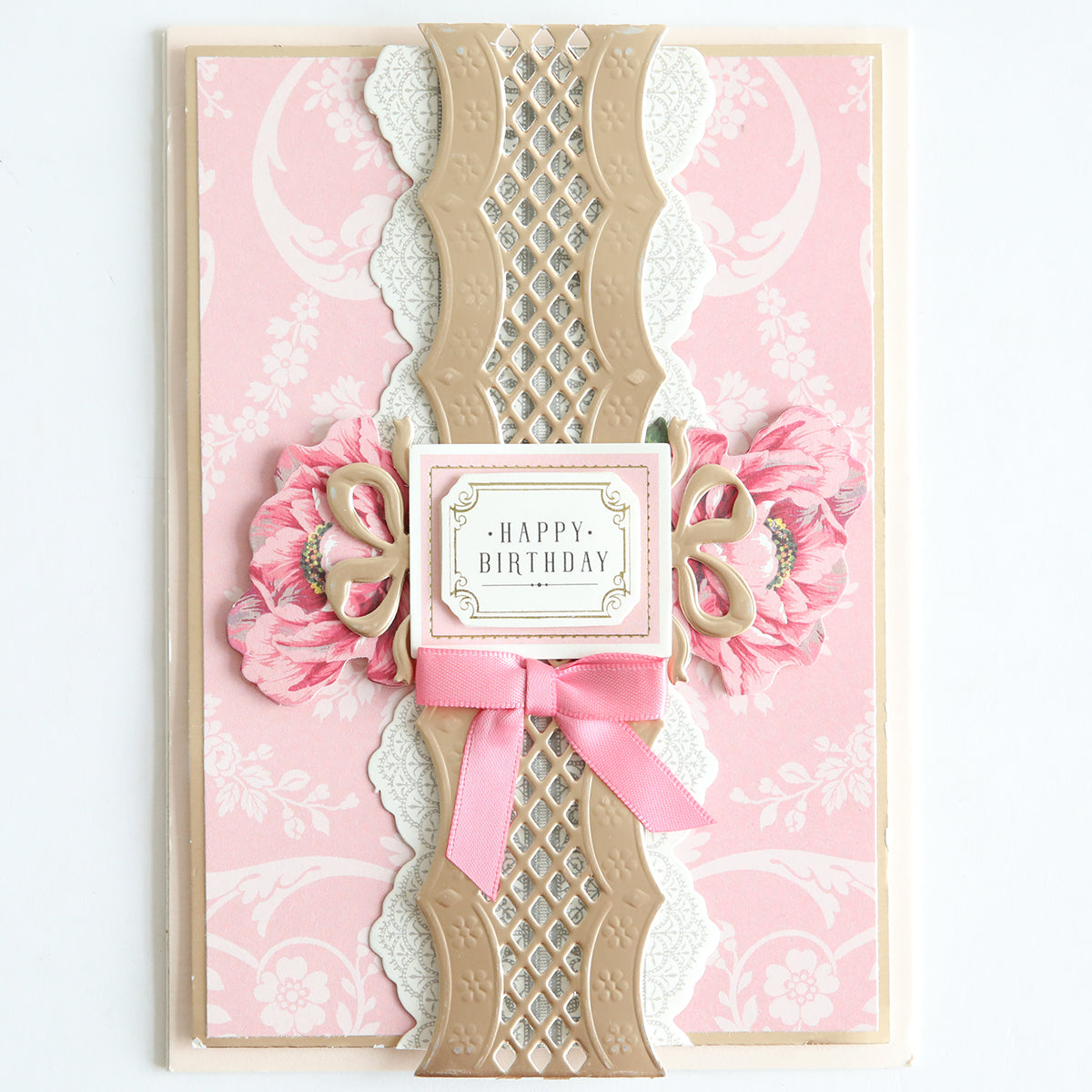 A birthday card crafted with the 12″ Regal Trimmings Border Dies and Rose Gold Matte Foil Cardstock, featuring pink and beige floral patterns, layered decorative accents, a pink bow, and a “Happy Birthday” message in the center.