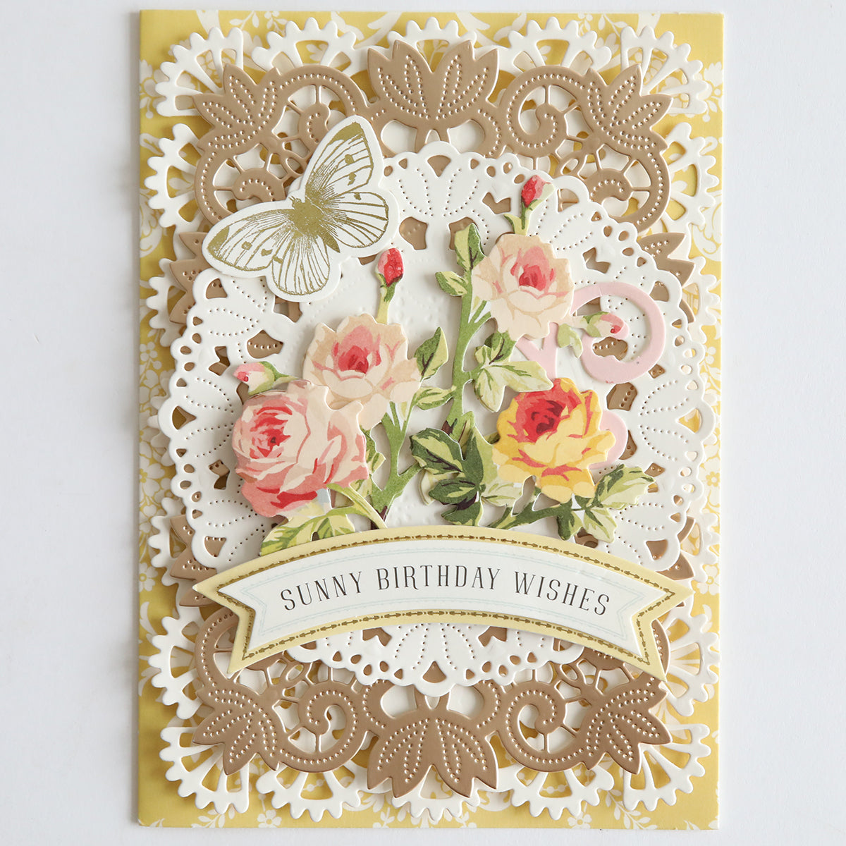 Celebrate a birthday with a decorative card crafted using the 12″ Regal Trimmings Border Dies and Rose Gold Matte Foil Cardstock, featuring layered lace borders, pink and yellow roses, an elegant gold butterfly, and the message “Sunny Birthday Wishes” on a banner at the bottom.