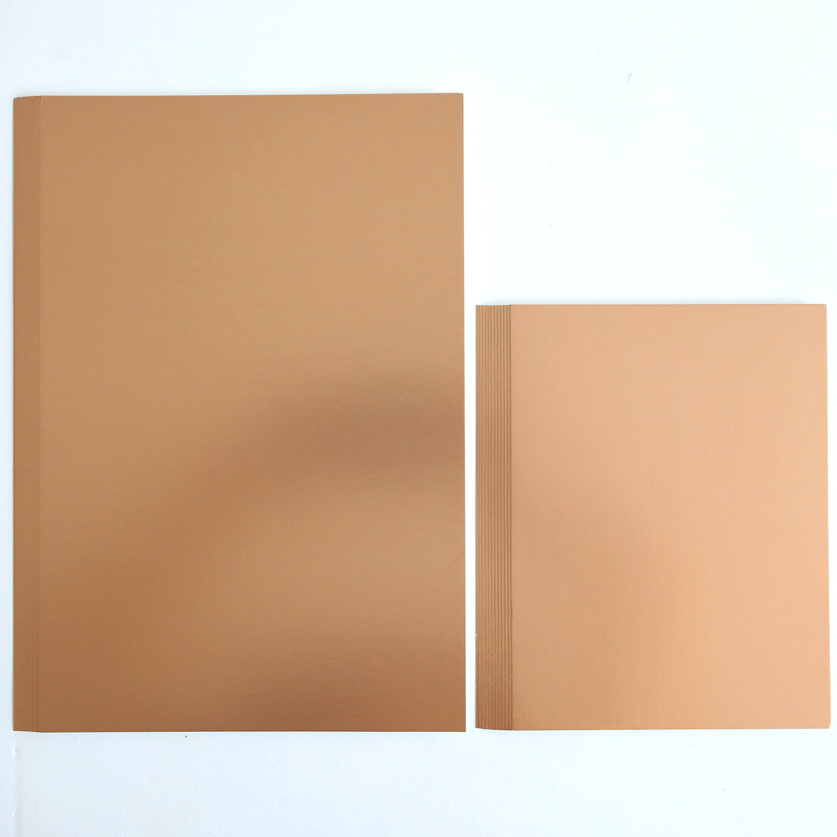 Two brown, blank softcover notebooks made from Luxury Matte Foil Cardstock Rose Gold rest side by side on a white surface—one larger, one smaller with visible page edges—perfect for diecutting or embossing projects.