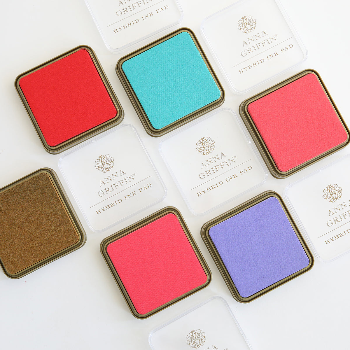 A collection of square ink pads from the Smitten Hybrid Ink Palette, featuring a range of colors such as red, teal, and purple, are arranged on a white surface. This selection includes both metallic and hybrid options. The packaging prominently showcases the brand name "Anna Griffin.