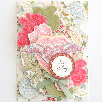 Valentine's Day card adorned with layered pink flowers and a lace-inspired background, enhanced using the Smitten Hybrid Ink Palette. The design is completed with a heart bearing wings and a green bow, featuring a round emblem with the message "Be Mine Valentine.