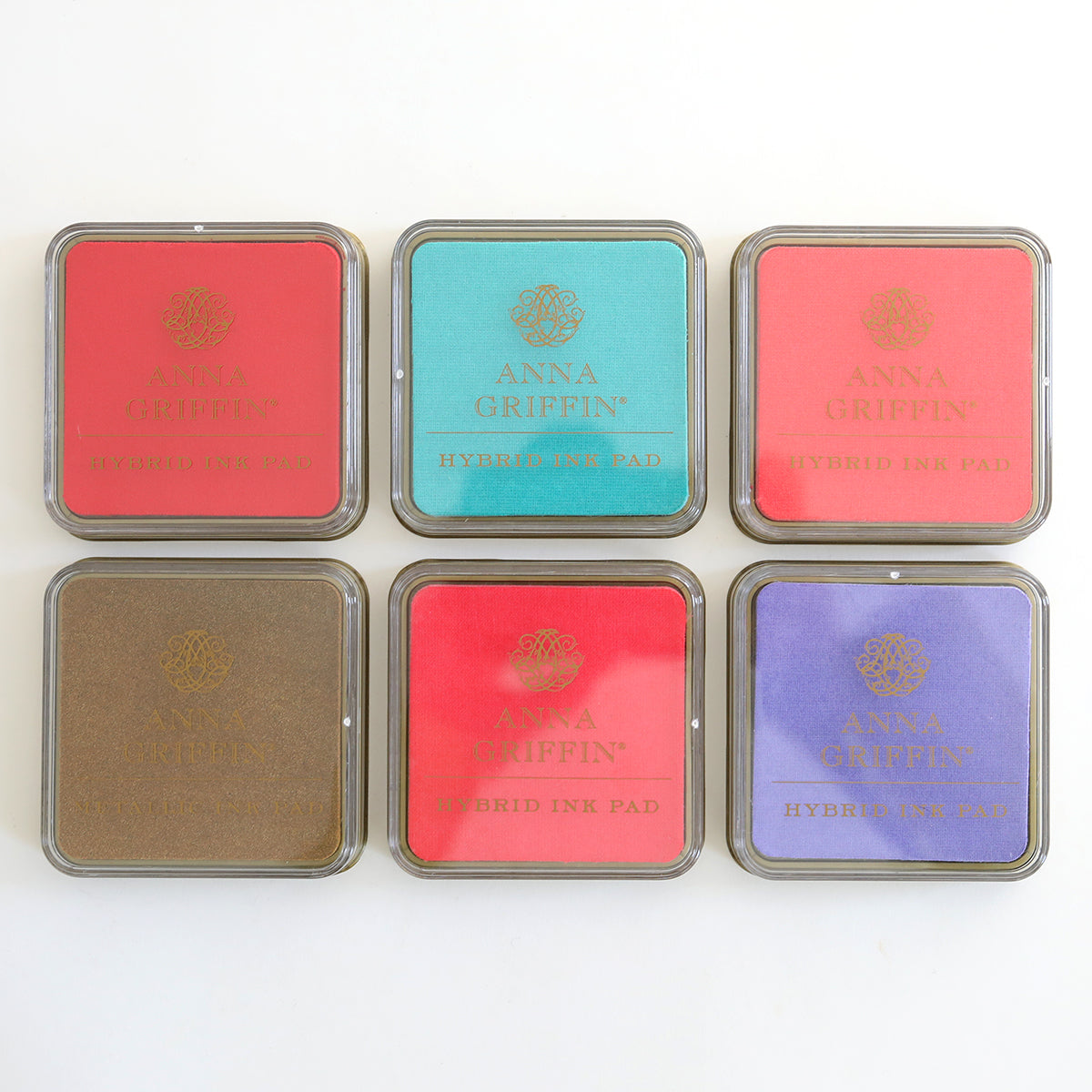 Six square ink pads from the Anna Griffin Smitten Hybrid Ink Palette, showcasing hues of red, teal, pink, brown, coral, and purple, are displayed in two rows on a white background.