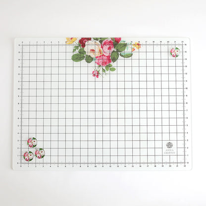 The Glass Magnetic Craft Mat 16 x 22 features a grid pattern, floral designs in the corners, and a logo in the bottom right. Ideal for precise cutting and crafting projects.