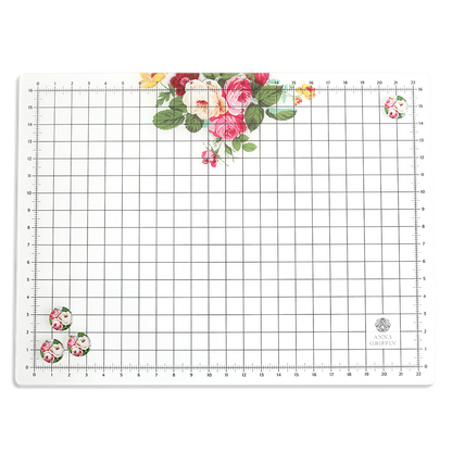 The Glass Magnetic Craft Mat 16 x 22 is a rectangular mat with grid and ruler markings, featuring clusters of pink, red, and yellow flowers in two corners and the "Anna Griffin" logo in the bottom right.