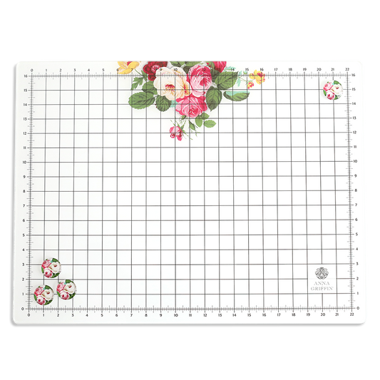 The Glass Magnetic Craft Mat 16 x 22 is a rectangular mat with grid and ruler markings, featuring clusters of pink, red, and yellow flowers in two corners and the "Anna Griffin" logo in the bottom right.