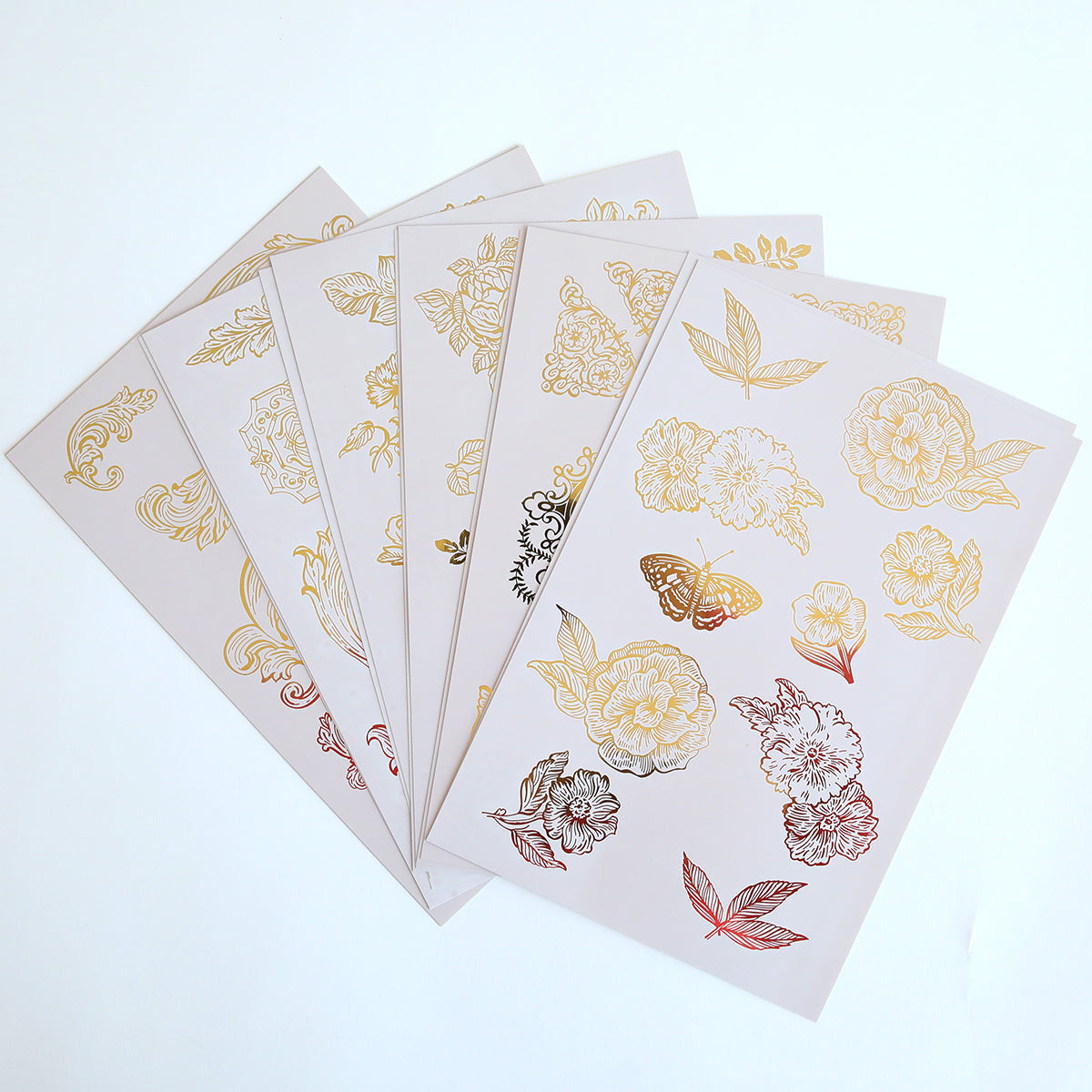 Enhance your crafts with Glorious Foil Rub Ons Gold, featuring five sheets of floral and butterfly designs with gold and black outlines on a light background. These gold foil transfers add elegance and create stunning effects.