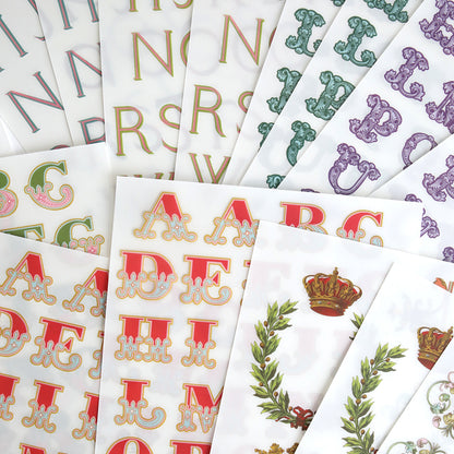 Fancy Alphabet Rub On Transfers, ideal for scrapbooking and cardmaking, come in a range of colors and ornate styles, with one sheet featuring a crown and laurel wreath design.