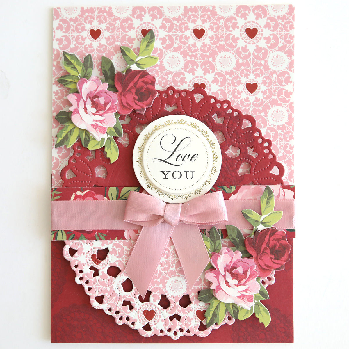 Crafted from Heartfelt Cardstock, this decorative card features pink and red florals, a pink ribbon bow, heart cutouts, and “Love You” in a gold-rimmed circle at the center.
