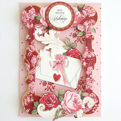 A decorative Valentine’s Day card made from Heartfelt Cardstock features pink and red floral designs, an envelope with a pink bow, and a “Be Mine Valentine” circular label.
