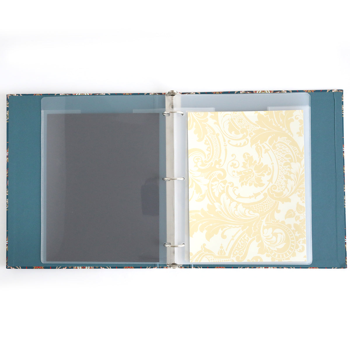 The Jasmine Die Storage Binder, featuring teal inner covers, is ideal for craft organization and includes two transparent sleeves with patterned paper sheets—one dark and one light with a floral design.