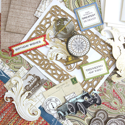 A flat lay of the All About Him Papercrafting Kit with Dies, featuring greeting cards, postcards, a compass, globe, and ornate paper cutouts on a paisley fabric background.