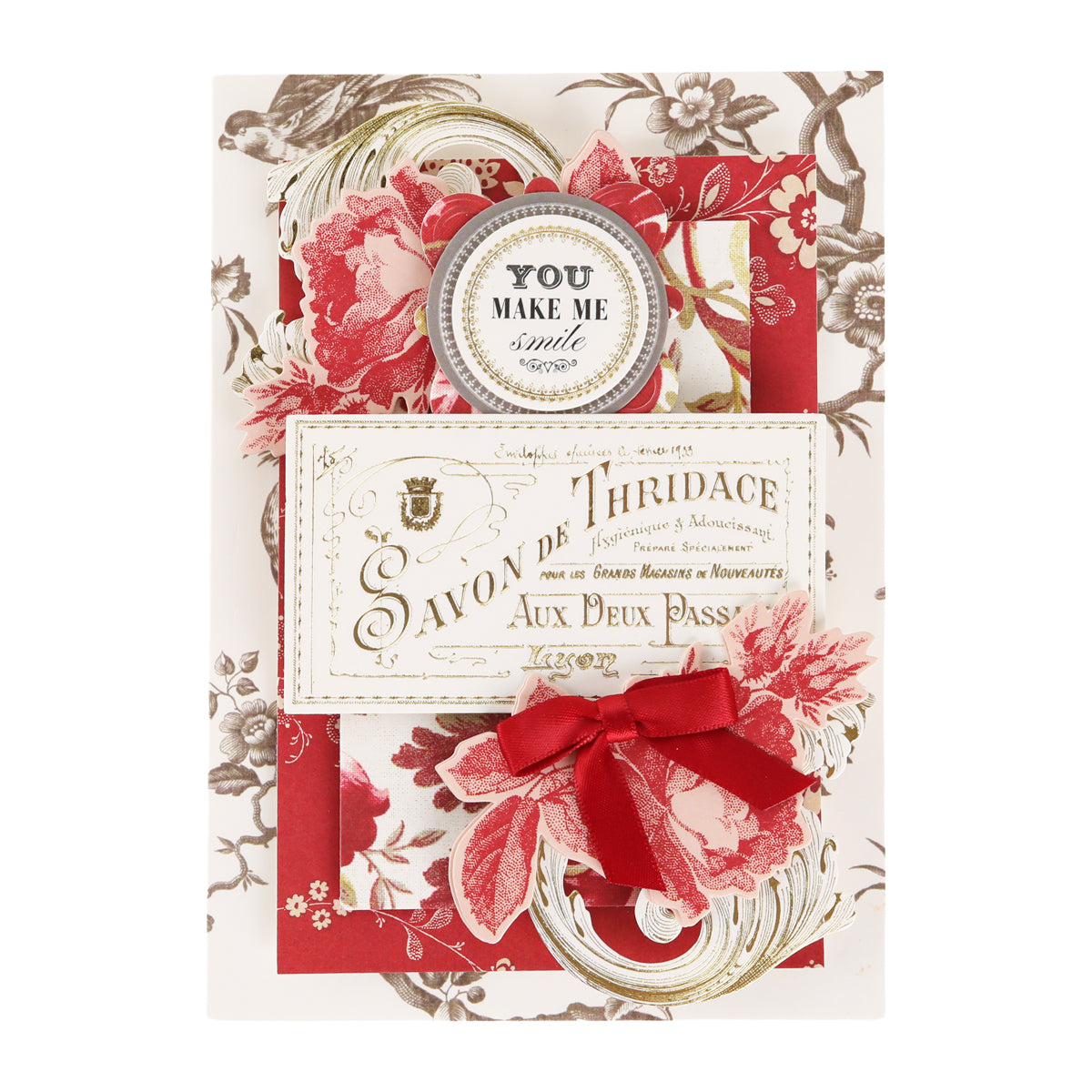 Greeting card featuring red floral accents, crafted using the Fleur Rouge Papercrafting Kit with Dies, a vibrant red ribbon, and a circular badge reading “YOU MAKE ME smile.” Vintage French text and ornamental details make it an ideal project for enthusiasts of vintage-inspired papercrafting and elegant die-cut designs.