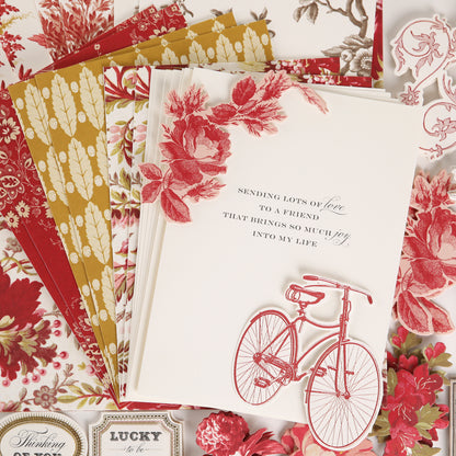 A stack of cards and papers from the Fleur Rouge Papercrafting Kit with Dies showcases vintage-inspired floral designs, with the top card featuring red flowers, a red bicycle, and a printed friendship message in black text.