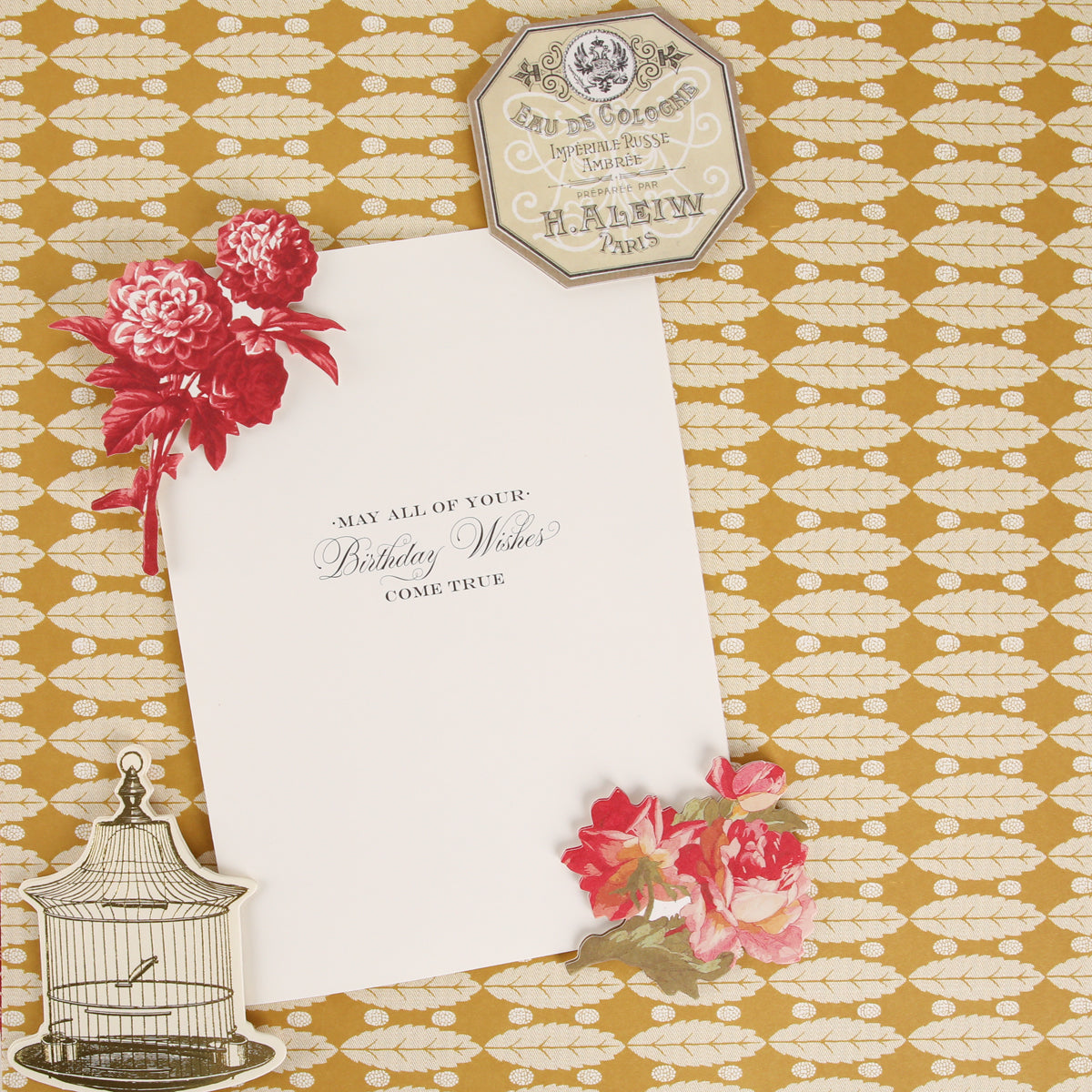 A white card with birthday wishes rests on a gold patterned surface, adorned with red flowers, a birdcage illustration, and a vintage hexagonal Paris label—crafted using the Fleur Rouge Papercrafting Kit with Dies, perfect for adding classic charm to your vintage-inspired papercrafting projects.