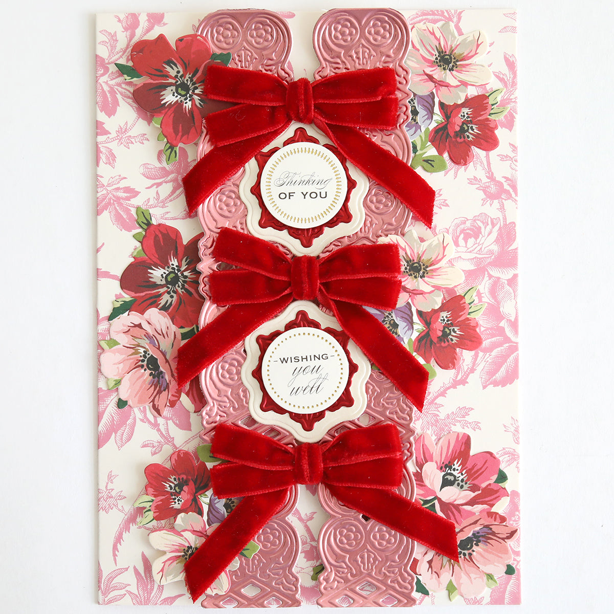 Three ornate pink Velvet Double Bows with floral tags rest on a pink and white flower-patterned background—perfect for holiday crafts or card making.