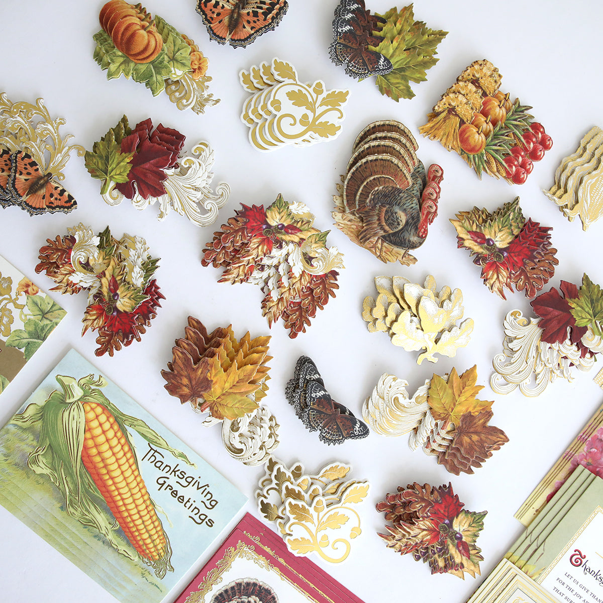The Fall Thanksgiving Postcards and Embellishments set features an assortment of autumn-themed paper cutouts—including turkeys, leaves, butterflies, and vegetables—arranged on a white surface with greeting cards in the corners. This collection is perfect for creating festive autumn paper crafts or adding a charming seasonal touch to your Thanksgiving postcards.