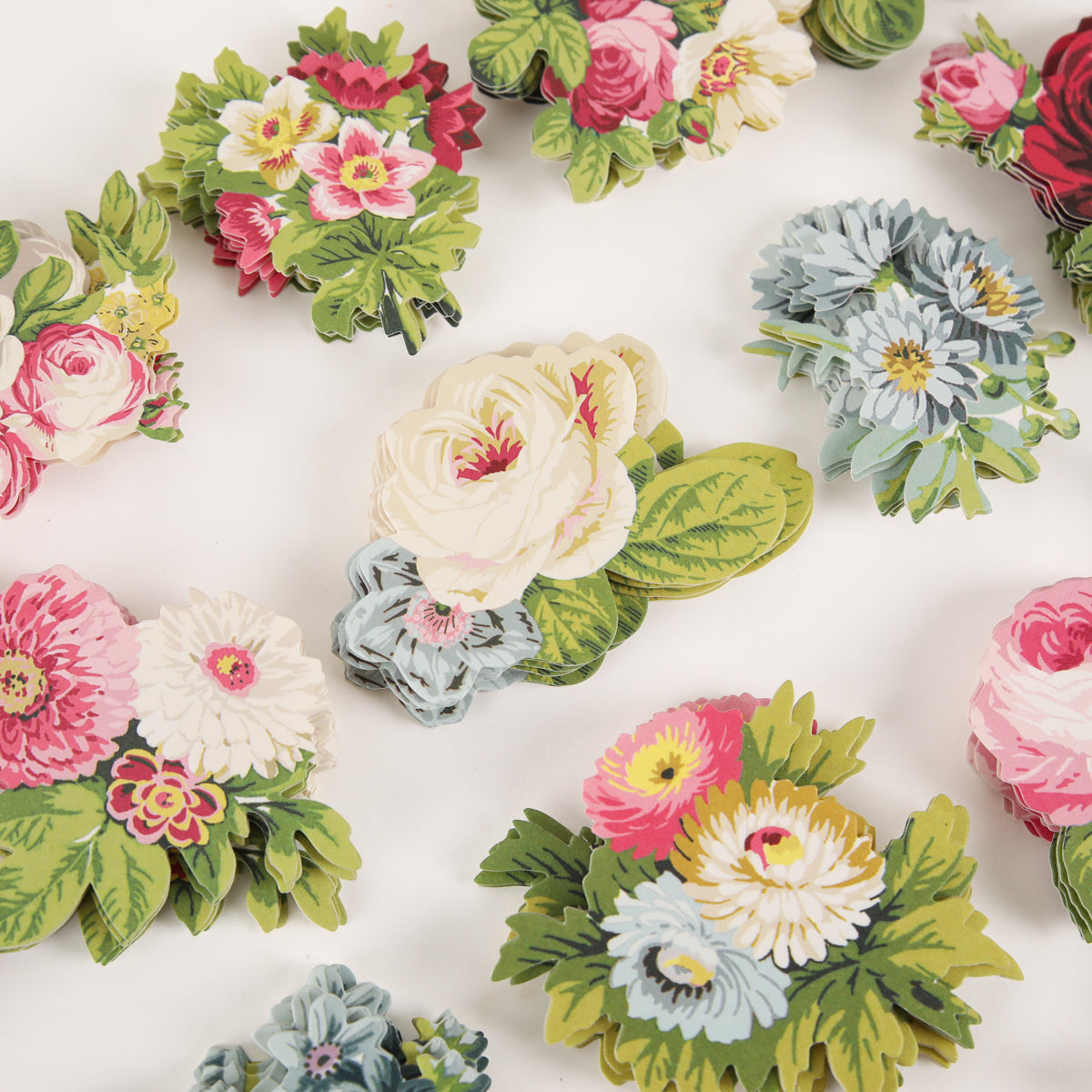 Several paper cutouts of colorful flower bouquets, reminiscent of the delightful Cottage Roses Embellishments, are arranged on a white surface. These vibrant embellishments feature roses, daisies, and chrysanthemums in pink, white, blue, and yellow hues accented with lush green leaves.