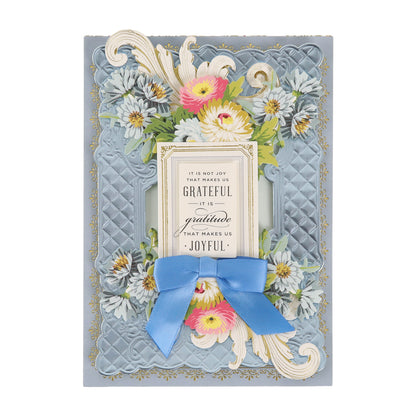 A decorative card adorned with floral designs and an elegant blue ribbon features the heartfelt message: “It is not joy that makes us grateful, it is gratitude that makes us joyful.” Cottage Roses Embellishments add a charming touch, making this card perfect for any special occasion.