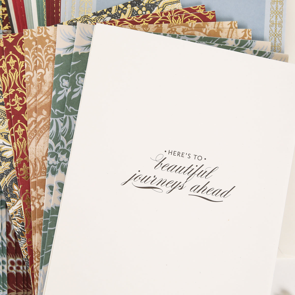 A stack of decorative cardstock from the Venetian Ultimate Paper Crafting Collection sits beneath a white card that says, “Here’s to: beautiful journeys ahead” in mixed fonts.