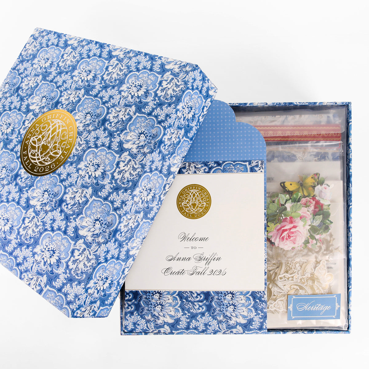 Introducing the Anna Griffin | Create Fall 2026: a beautifully designed blue patterned gift box with a gold seal, featuring an envelope, a welcome card, and a bag filled with decorative craft items like flowers and butterflies—ideal for new members joining our creative community or enhancing your online craft classes.