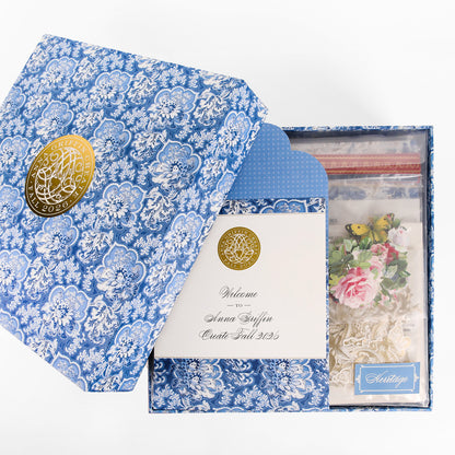 Introducing the Anna Griffin | Create Fall 2026: a beautifully designed blue patterned gift box with a gold seal, featuring an envelope, a welcome card, and a bag filled with decorative craft items like flowers and butterflies—ideal for new members joining our creative community or enhancing your online craft classes.