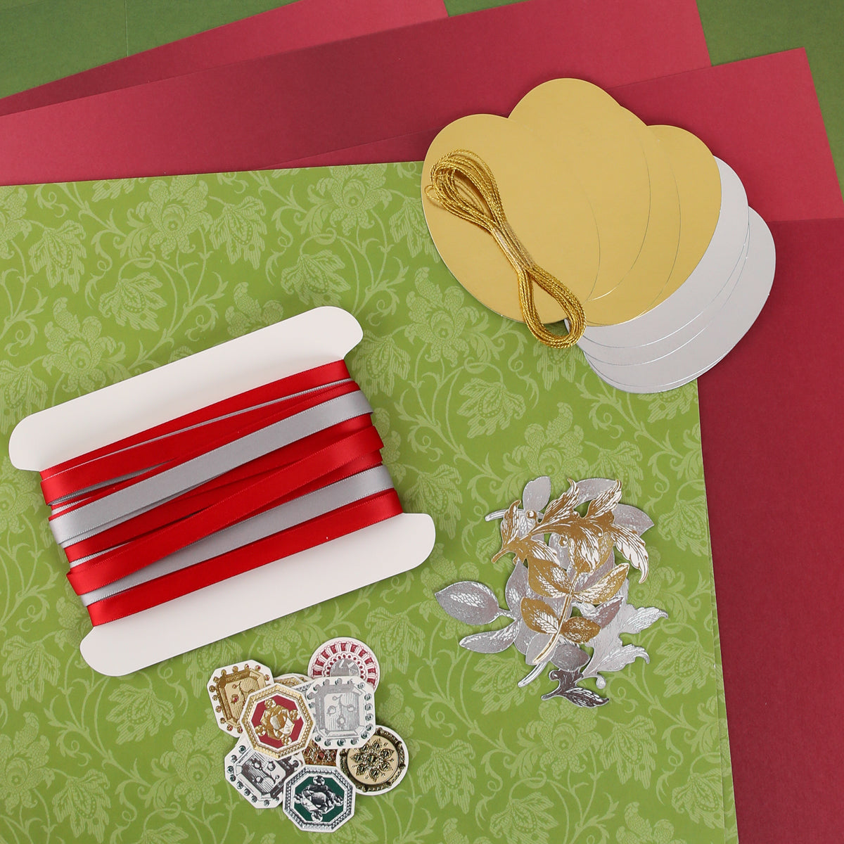 A variety of craft materials—including red and silver ribbons, gold and silver tags, gold thread, stickers, and patterned paper—are arranged on a flat surface, offering perfect inspiration for creative classes or community gatherings centered around the Anna Griffin | Create Fall 2026 collection.