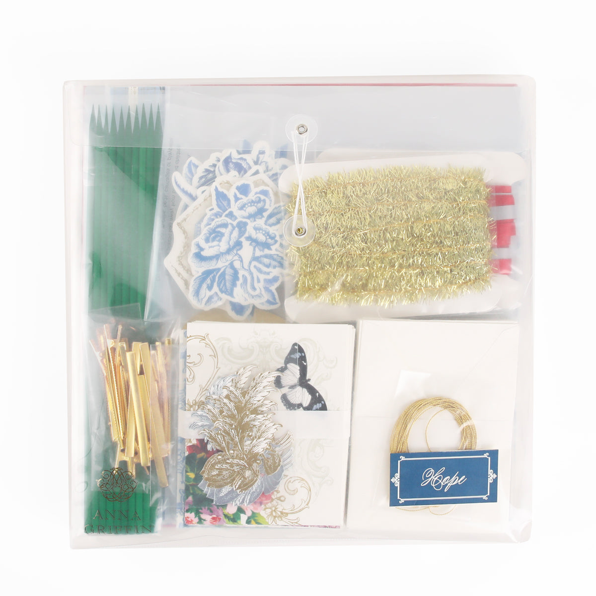 A clear pouch containing craft supplies—gold string, paper cutouts, gold tinsel trim, green sticks, envelopes, and decorative cards with floral and butterfly designs—from Anna Griffin | Create Fall 2026; perfect for heritage crafts or as part of your crafting classes.