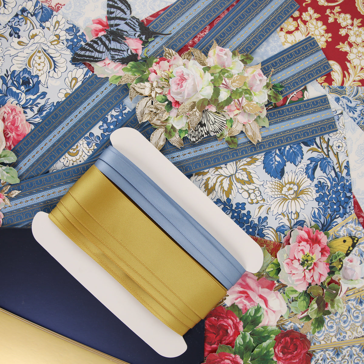 Gold and blue ribbons on a white spool rest atop floral patterned papers featuring blue, red, and gold designs with flowers and butterflies—offering perfect inspiration for the creative community and upcoming craft classes as you get ready for Anna Griffin | Create Fall 2026.