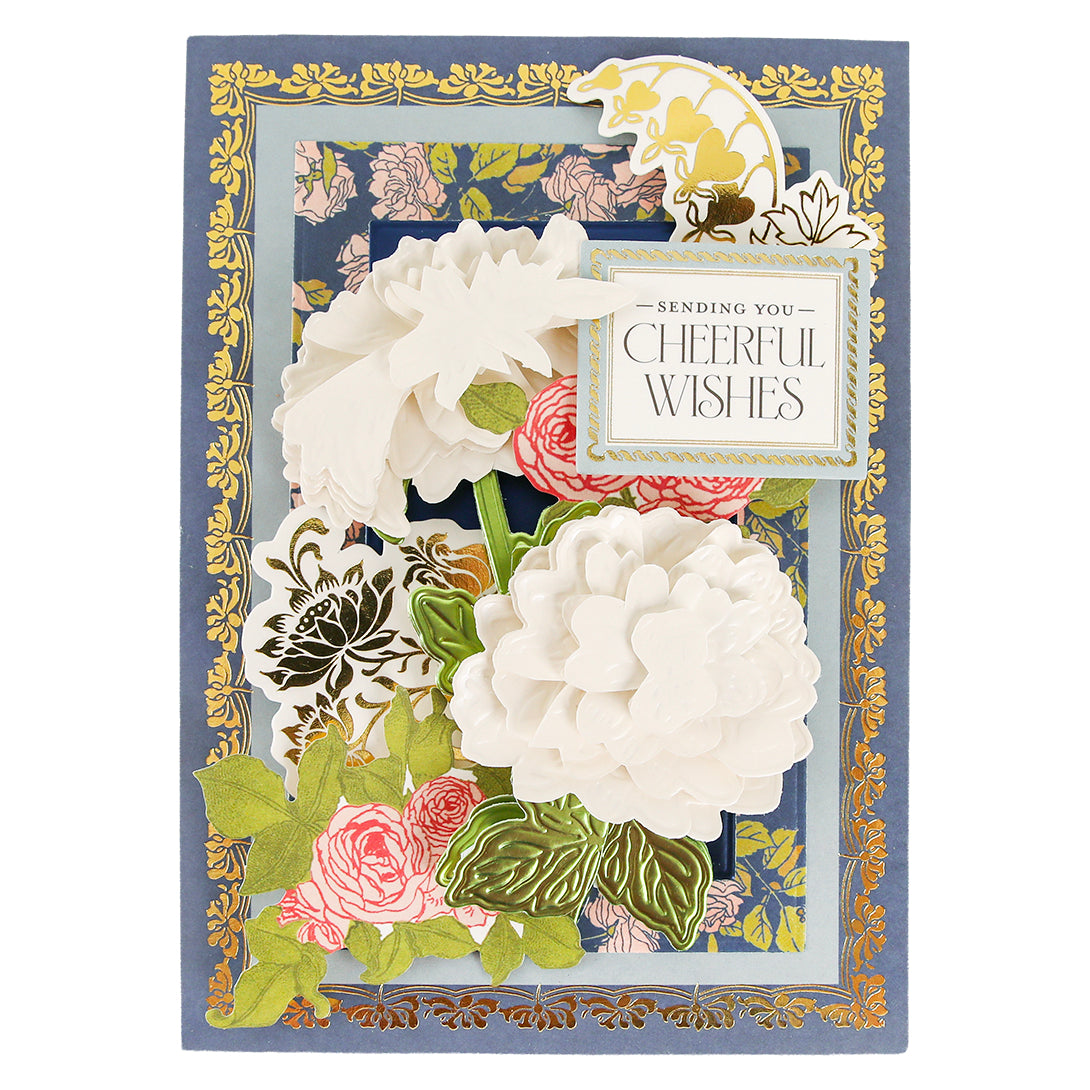 Greeting card featuring layered white and pink flowers crafted with the Dahlia, Mum, and Sunflower 3D Floral Dies, accented by green leaves, a gold floral border, and a sign reading “Sending You Cheerful Wishes” on a blue background—a charming celebration of paper crafts and botanical detail.