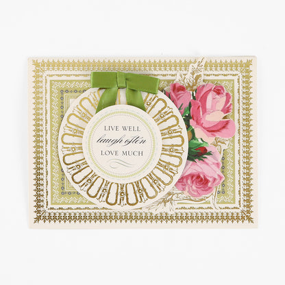 Certainly! Here’s a revised version using the provided product name: Greeting card featuring a gold decorative border, pink roses, and a round tag with green ribbon that reads “Live well, laugh often, love much.” From our Pretty Pattern A2 Cards and Envelopes collection, these handmade cards are paired with elegant ivory envelopes.