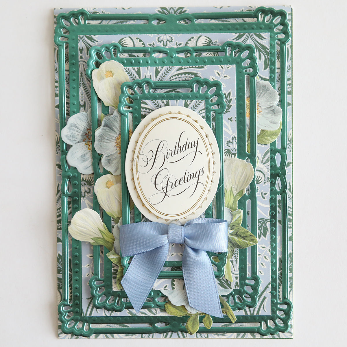 Elegant birthday card with a floral motif, featuring layered frames and a light blue bow on 12" x 12" sheets. It presents "Birthday Greetings" in a central oval, enhanced by the use of Paris Green foil matte cardstock for a sophisticated finish.