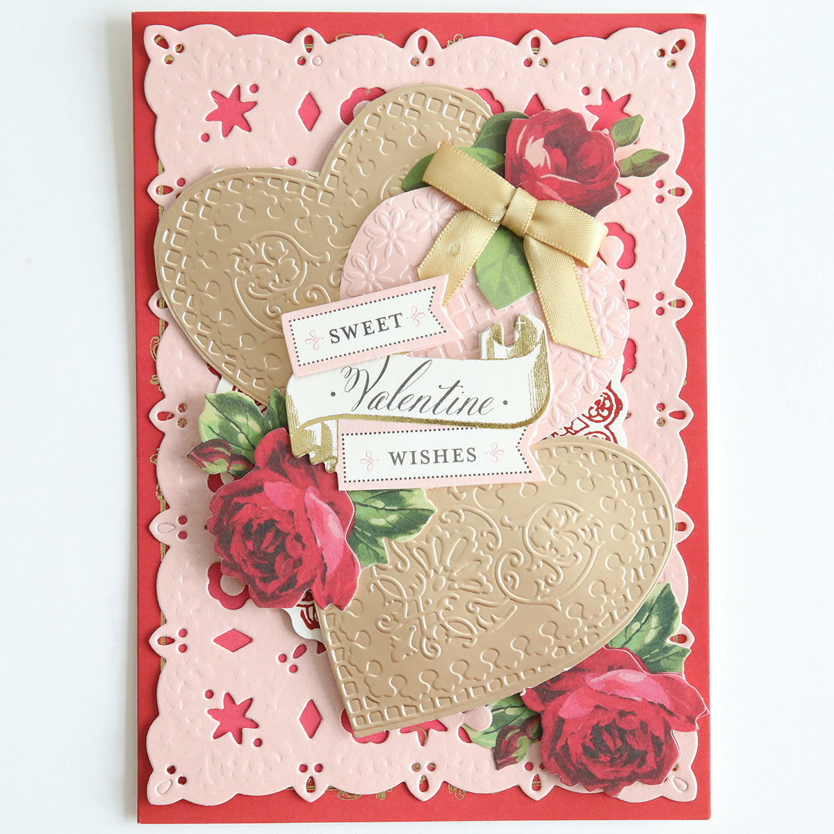 A decorative Valentine's Day card crafted from Foil Matte Cardstock in rose gold showcases layered hearts, red roses, and a gold bow. It features embossed patterns and elegantly displays the text "Sweet Valentine Wishes" on the front.