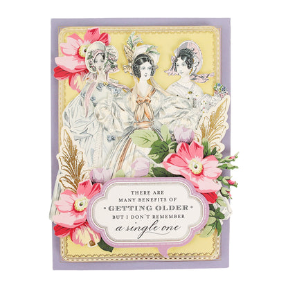 Three women, styled as Victorian ladies in vintage clothing, stand among colorful flowers with a sign reading, “There are many benefits of getting older but I don’t remember a single one.” The charming scene resembles the layered die cuts featured in the Paper Doll Card Toppers Kit, perfect for creating elegant greeting cards.
