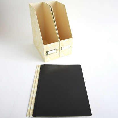 Two ivory damask die storage boxes stand upright, while three black oversized die storage boxes are lying flat in front of them on a white surface.